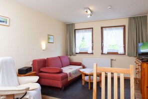 A furnished room with a red sofa, a small table, a wooden chair, a television, and two windows with curtains. - Vacation apartment 1 "Esche" - Haus Keller vacation apartments (Todtnau)