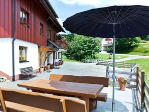 Un espace extérieur aménagé avec des meubles en bois, comprenant une table et des bancs, un parasol noir et un bâtiment rouge orné de jardinières.