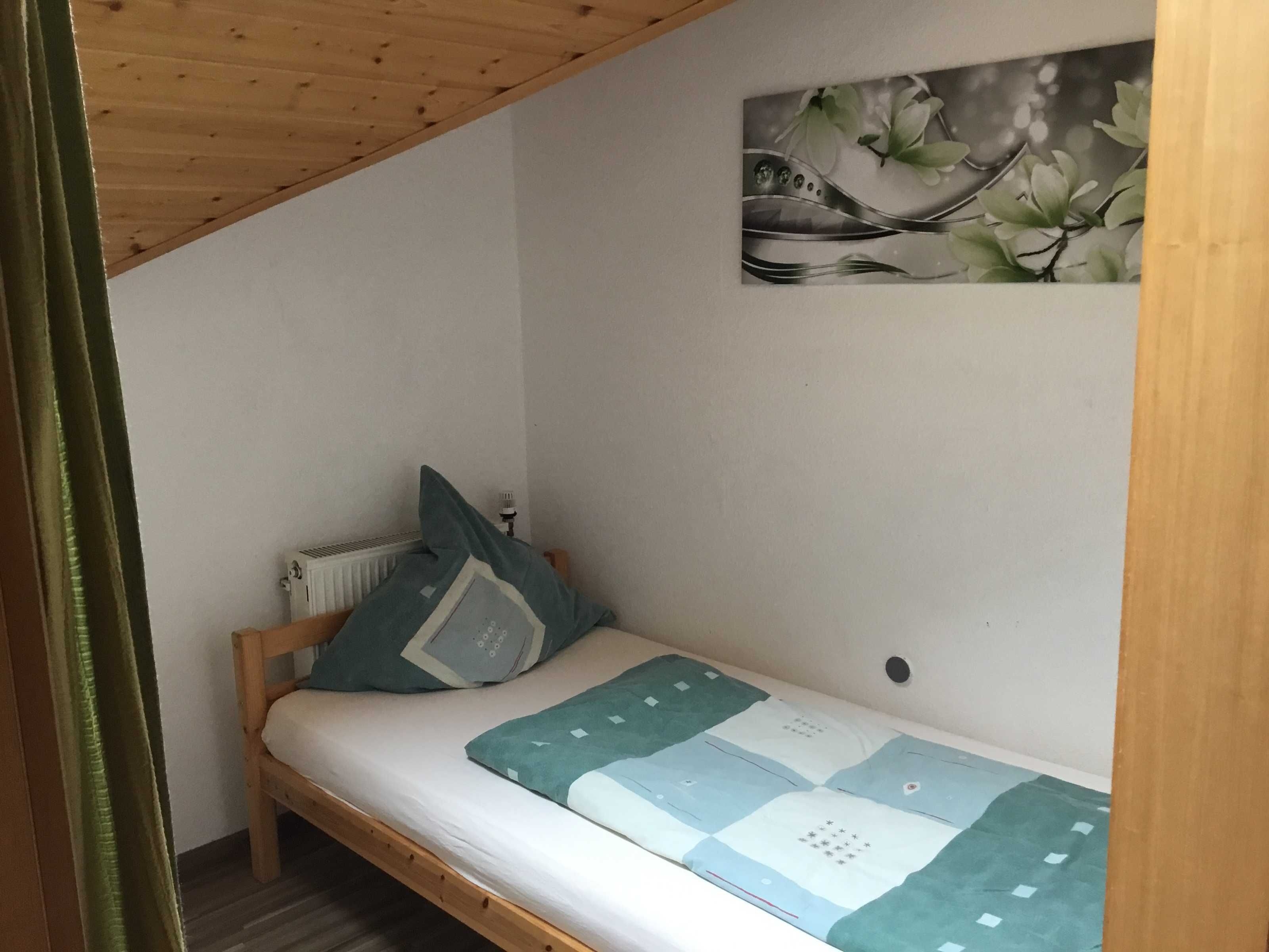 A small, single-bed room with a wooden ceiling and a radiator. A painting of flowers hangs on the wall.