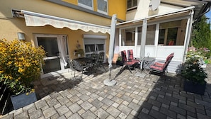 Un patio con mobiliario exterior, una zona de estar techada y un edificio amarillo con antena parabólica.