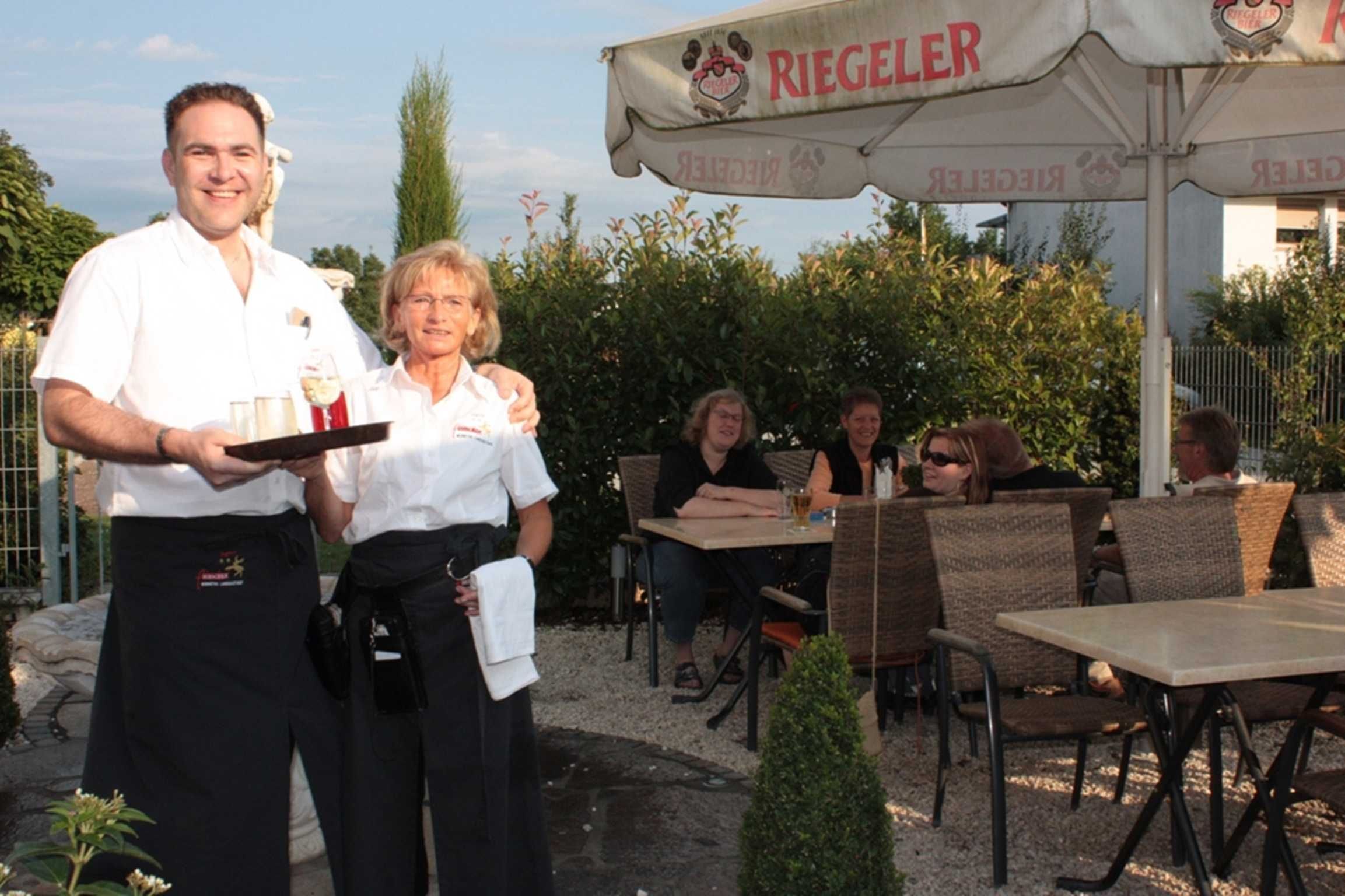 Un espace de restauration en plein air avec des chaises et des tables en osier, un serveur tenant un plateau et une banderole portant le nom « RIEGELER ».