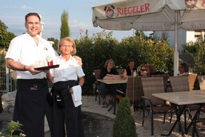 Un espacio de comedor al aire libre con sillas y mesas de mimbre, un camarero sosteniendo una bandeja y un cartel con el nombre 'RIEGELER'.