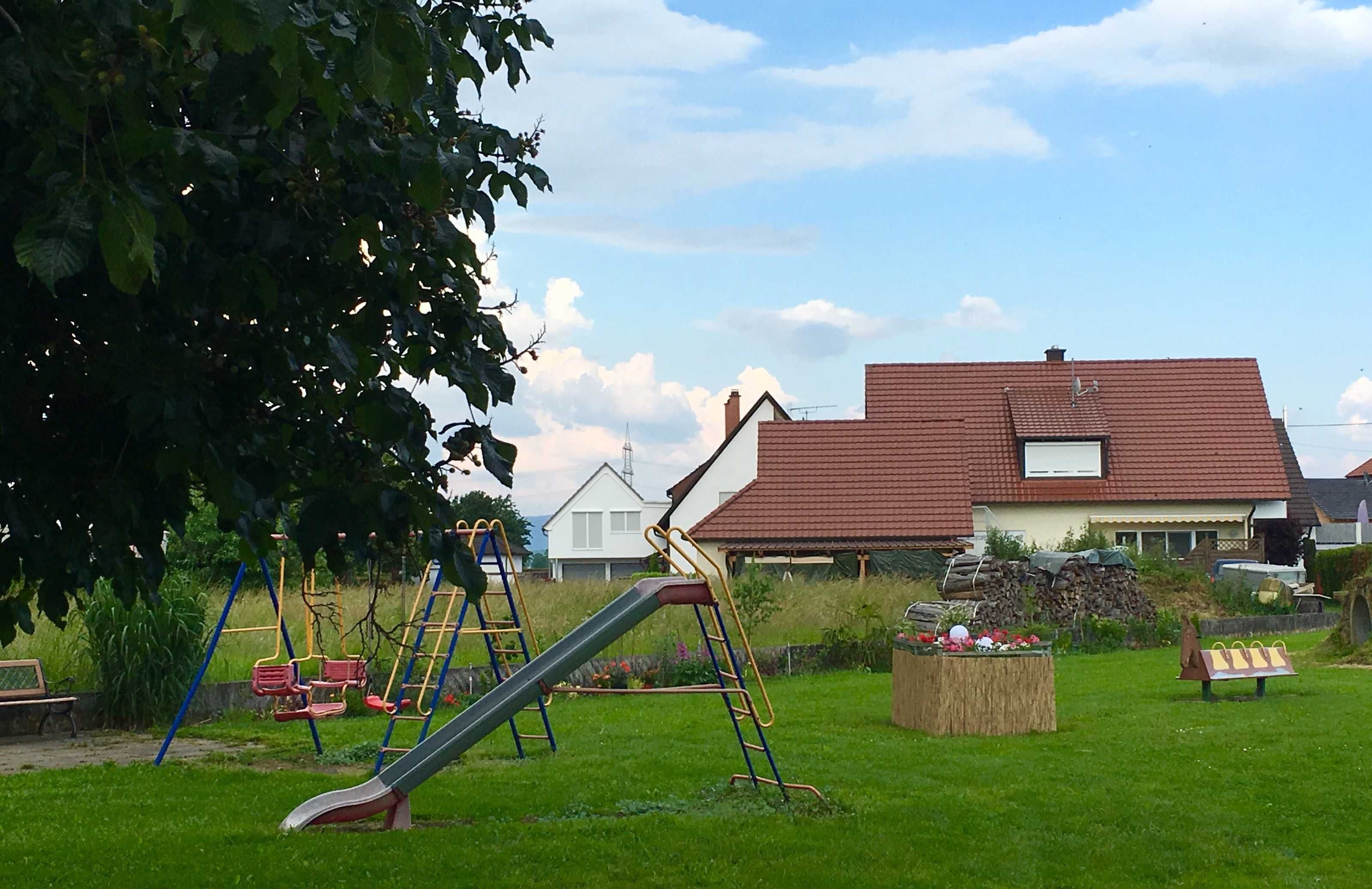 Une aire de jeux avec des toboggans et des balançoires, un banc en bois et, au fond, une maison avec un toit rouge.