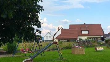 Une aire de jeux avec des toboggans et des balançoires, un banc en bois et, au fond, une maison avec un toit rouge.