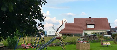 Un parque infantil con toboganes y columpios, un banco de madera y una casa con techo rojo al fondo.