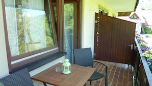 A small balcony with a wooden table and two chairs, a window with curtains, and a door with a wooden panel. - Vacation apartment Kapellenblick, 45sqm, 1 bedroom, max. 3 persons, 1 - 3 (Simonswald)