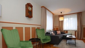 Un salon avec un canapé, deux fauteuils verts, une petite table, une horloge au mur et une fenêtre avec des rideaux.