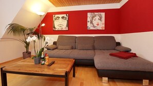Un salon avec un plafond en bois, un canapé gris, un mur rouge orné d’œuvres d’art, une table basse avec des boissons et une plante.