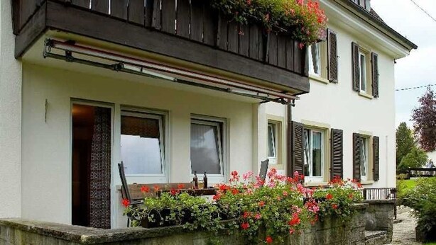 Une maison à deux étages avec un balcon, un mur en pierre et un jardin fleuri.