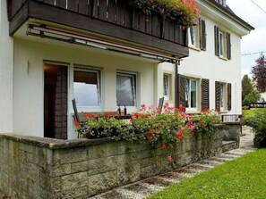 A two-story house with a balcony, a stone wall, and a garden with flowers.