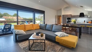 Ein modernes Wohnzimmer mit grauem Sofa, gelben Kissen und einem Couchtisch. Es gibt einen Essbereich mit Tisch und Stühlen sowie eine Küche mit einer Bar.