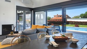Ein modernes Wohnzimmer mit einem großen Glasfenster, das zu einem Poolbereich führt, einem Kamin und einem Esstisch, der mit Schüsseln und einer Obstschale gedeckt ist.