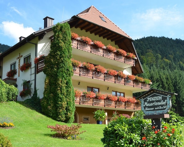 A multi-story building with balconies adorned with flowers, a sign indicating a pension, and a lush green garden. - Apartment Talblick - Mosertonihof (Elzach)