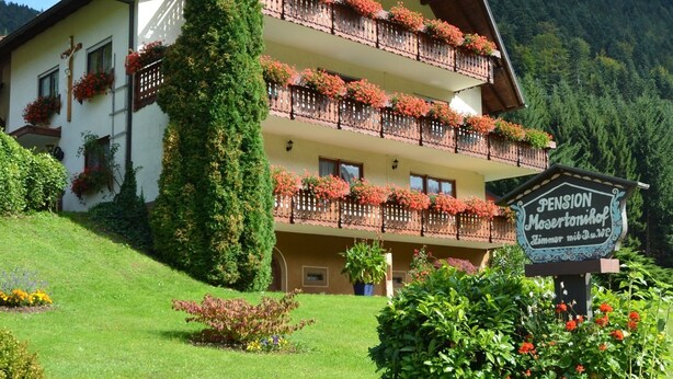 A multi-story building with balconies adorned with flowers, a sign indicating a pension, and a lush green garden.