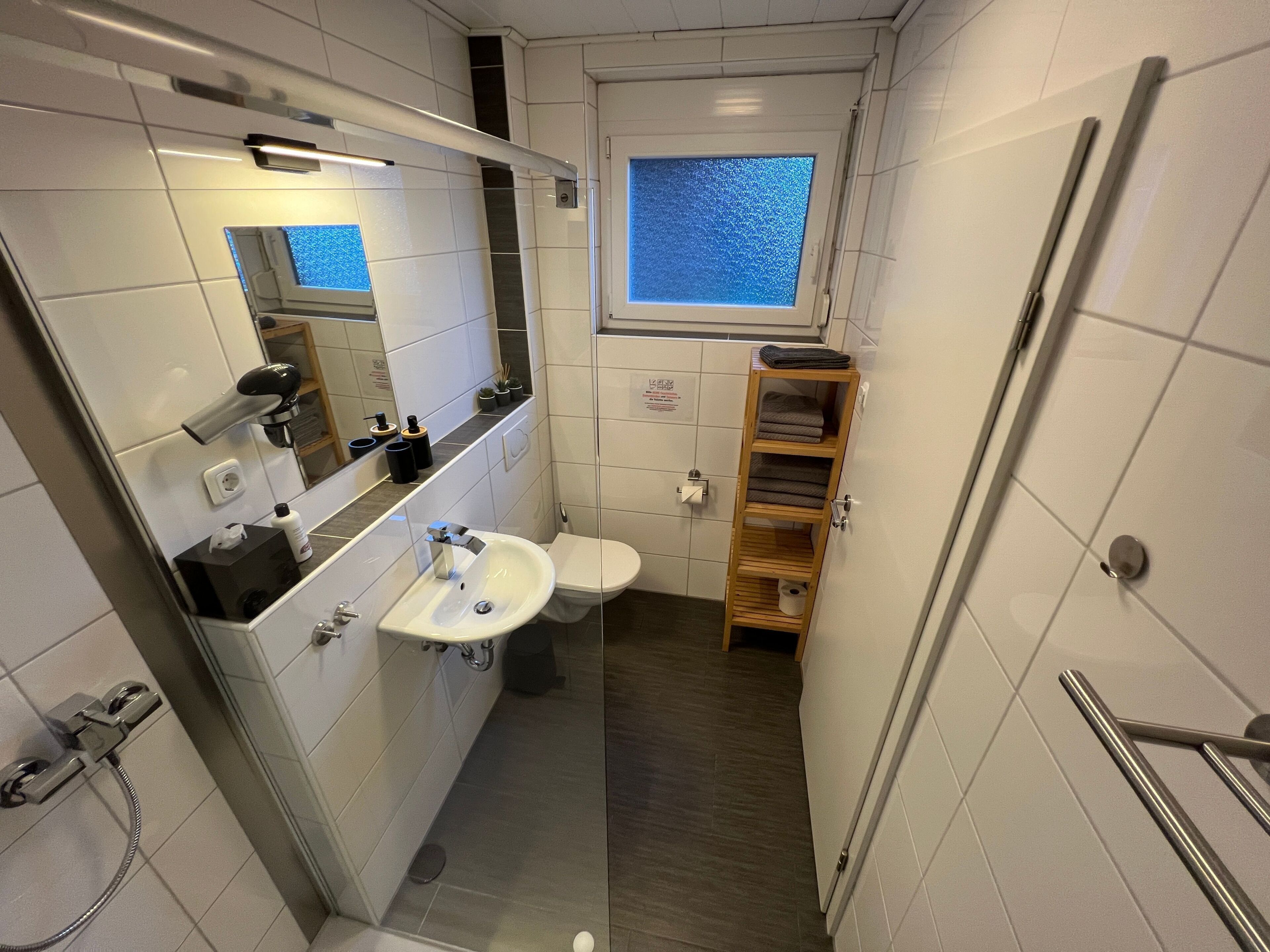 Ein kleines, modernes Badezimmer mit Waschbecken, Spiegel und Toilette in Weiß. Über der Toilette befindet sich ein Fenster, darüber hinaus ein Ablagebereich mit Handtüchern und ein Duschbereich mit Duschbrause.