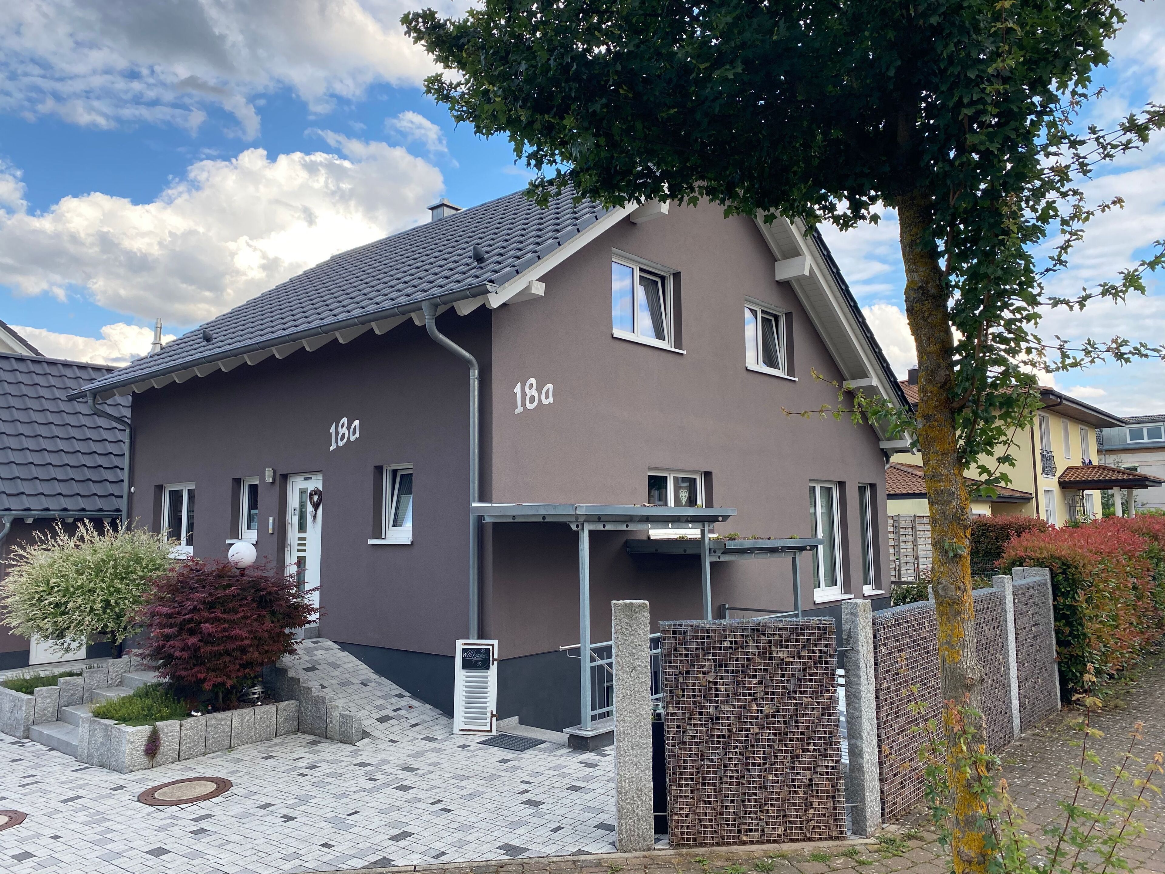 Ein zweistöckiges Gebäude mit der Hausnummer '18a', einem überdachten Eingang und einem kleinen Garten mit Stufen zum Eingang.