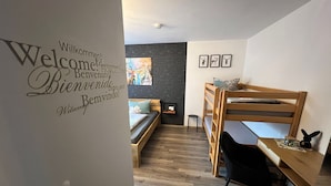 Ein Zimmer mit Etagenbett, kleinem Schreibtisch, Stuhl, Wandregal und einer Wand mit dekorativem Empfangstext in verschiedenen Sprachen.