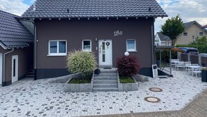 Ein zweistöckiges Gebäude mit dunkelbrauner Fassade, Hausnummer 18a, mit einem Ziegeldach, einer Haustür und einer kleinen Gartenanlage mit Pflanzen.