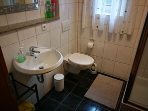 Une salle de bain avec un lavabo, des toilettes et une petite poubelle. Un miroir est placé au-dessus du lavabo, et on peut voir un porte-serviettes.