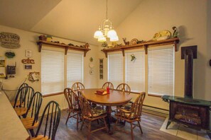 A dining area with a round table and six chairs, flanked by two windows with blinds, a hanging light fixture, and a fireplace.