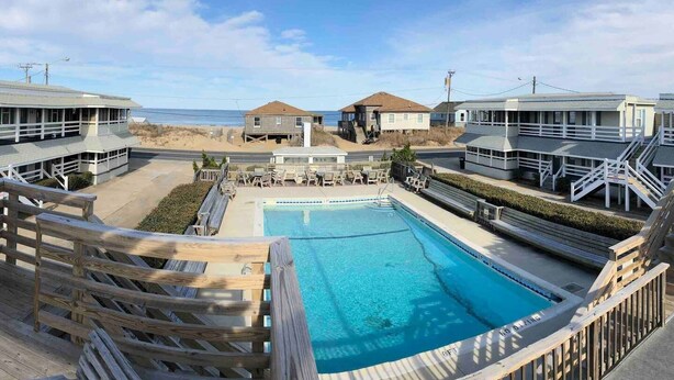 A pool area with a view of the beach and ocean, wooden deck chairs, and a lifeguard tower. - Saltaire Cottages14: Just Renovated! 4BR Private Beach Access Ocean View Pool! (Kitty Hawk)