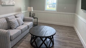 A living room with a striped sofa, a round coffee table, a lamp, and a window with a view of a car outside. - Saltaire Cottages14: Just Renovated! 4BR Private Beach Access Ocean View Pool! (Kitty Hawk)