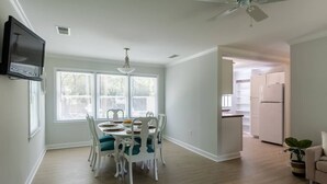 Un comedor con una mesa dispuesta para cuatro, sillas blancas, un ventilador de techo y un televisor montado en la pared.