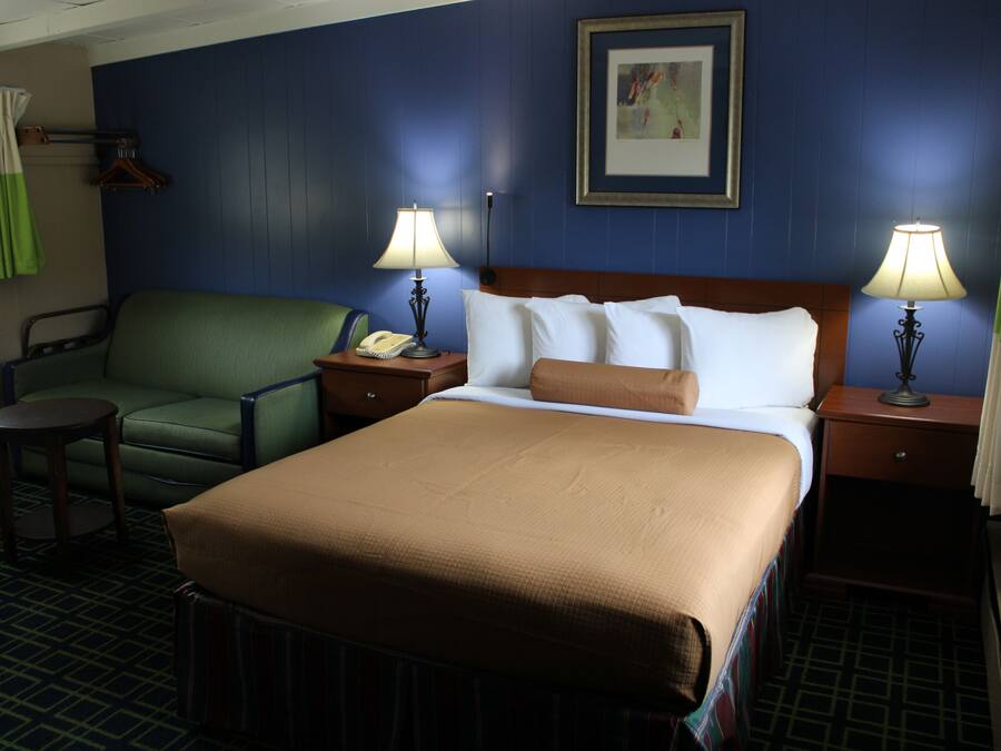 A hotel room with a large bed, two bedside tables with lamps, a green armchair, a small round table, and a framed picture on the wall.