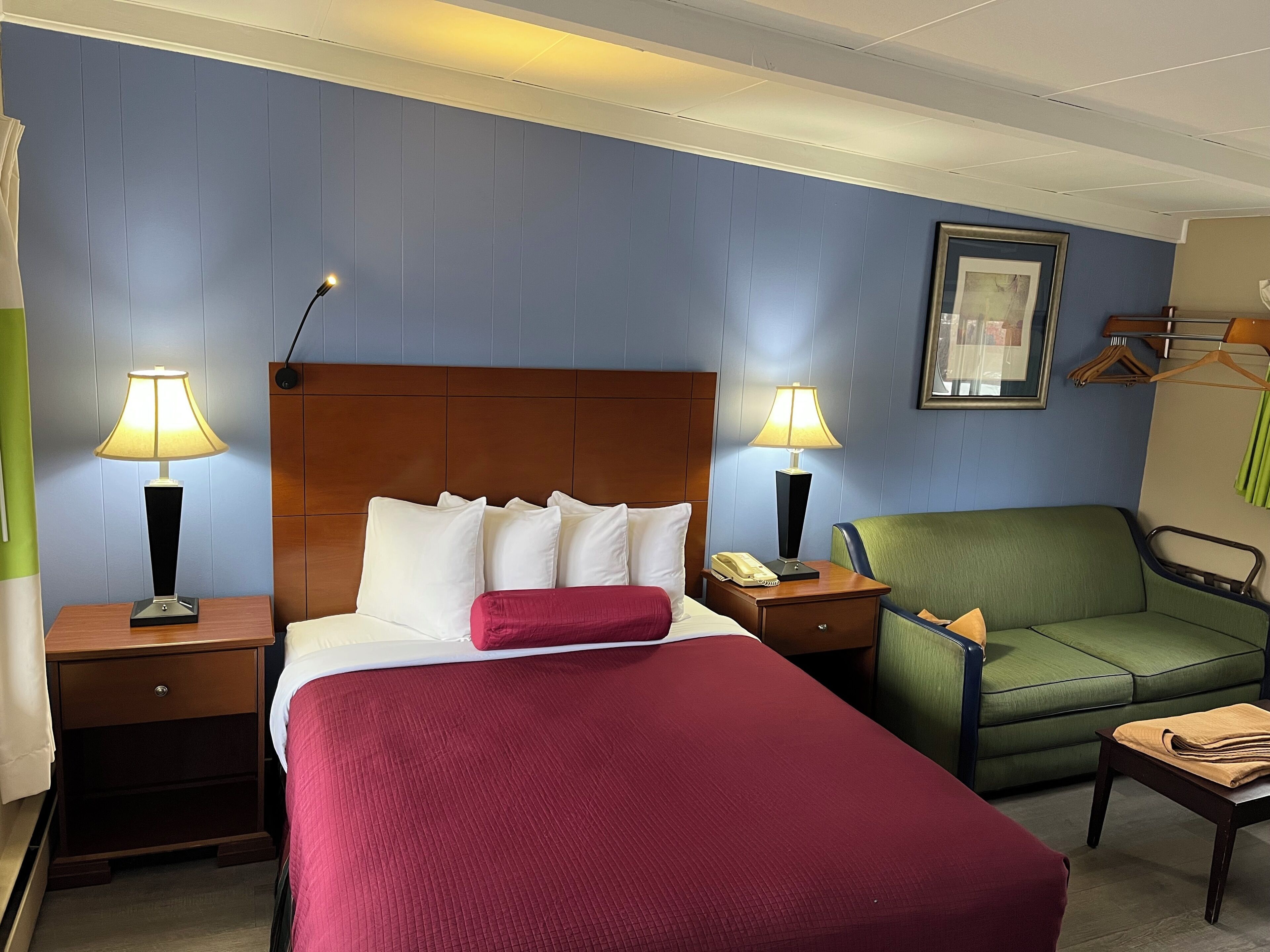 A hotel room with a bed, two bedside tables with lamps, a green armchair, a wooden nightstand, and a framed picture on the wall.