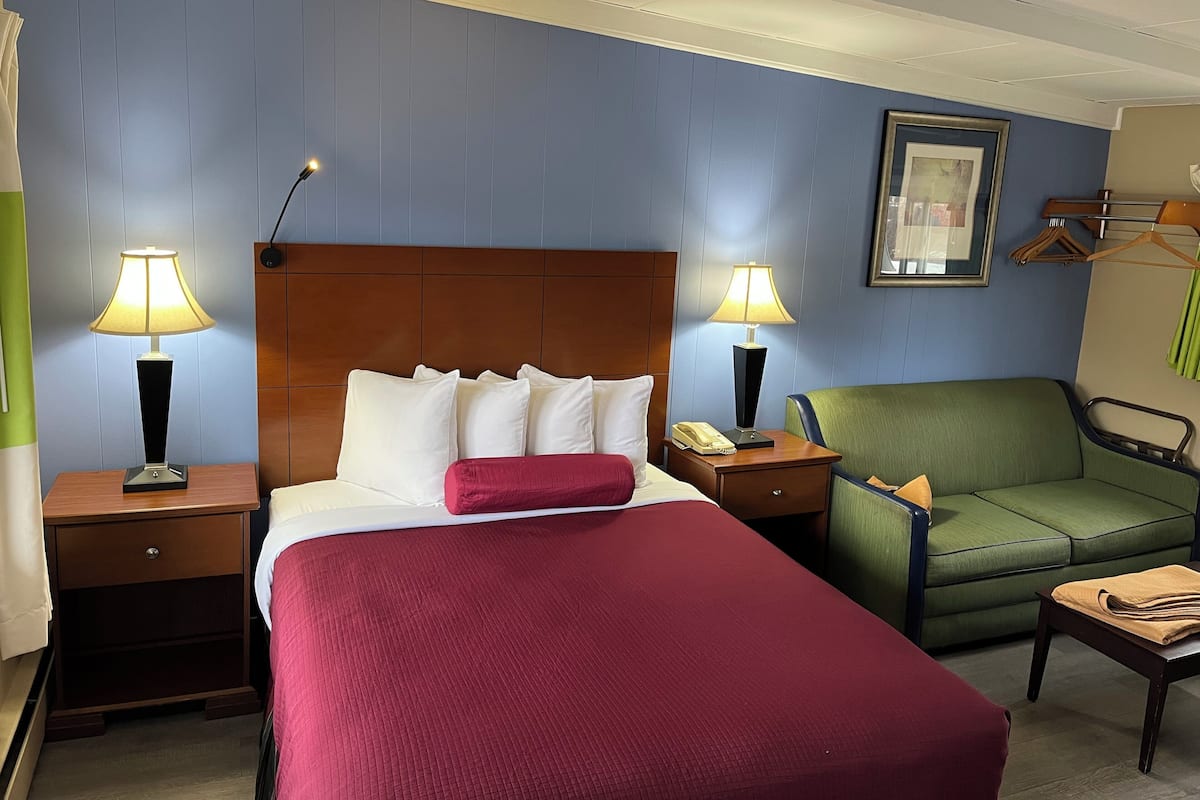 A hotel room with a bed, two bedside tables with lamps, a green armchair, a wooden nightstand, and a framed picture on the wall.