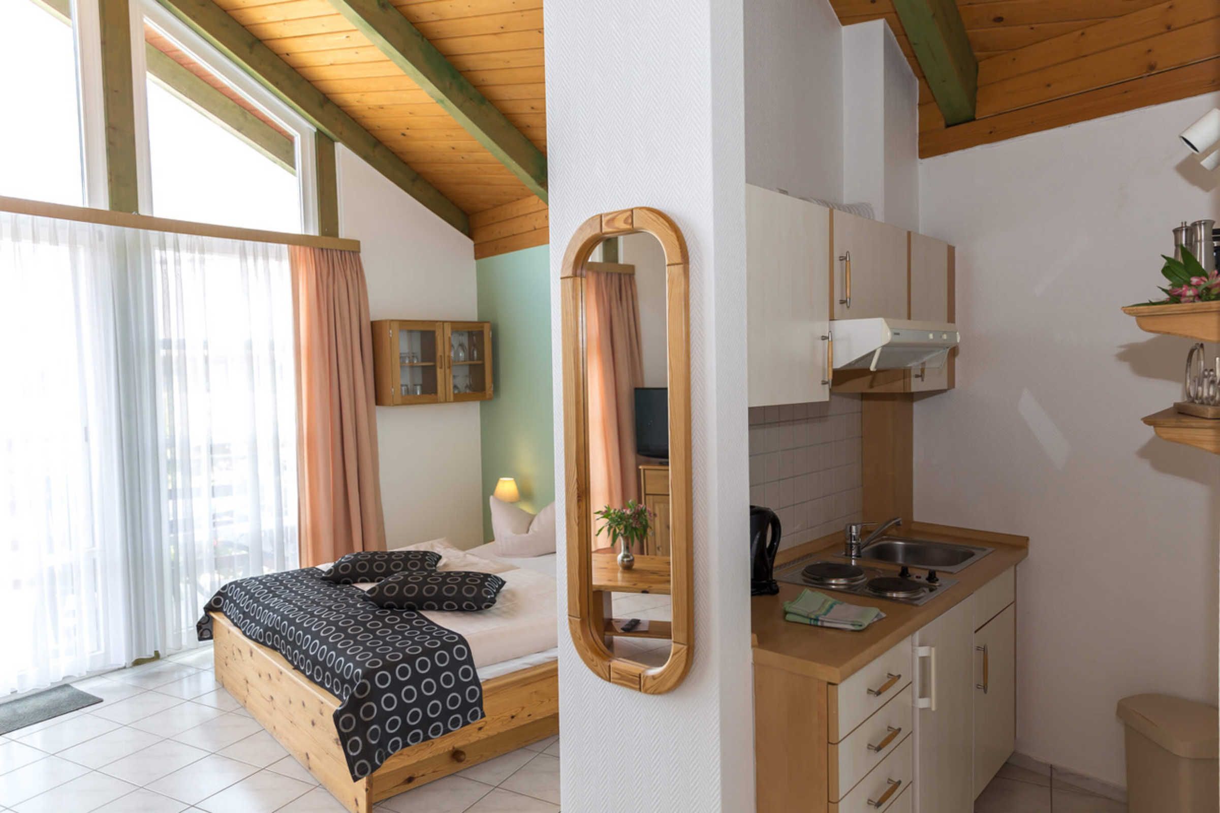 A bedroom with a bed, a bedside table, a mirror, and a window with curtains. The image also shows a kitchenette with a sink, a stove, and cabinets.