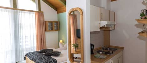 A bedroom with a bed, a bedside table, a mirror, and a window with curtains. The image also shows a kitchenette with a sink, a stove, and cabinets.