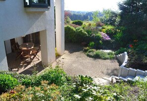 En terrasse med bord og stole, omgivet af en have med forskellige planter og en stenvæg.
