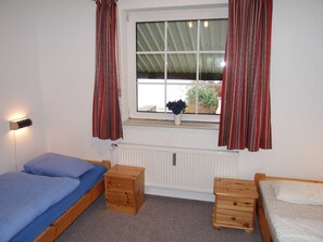Um pequeno alojamento de um único quarto, com uma cama, uma pequena mesa de cabeceira de madeira, um radiador e uma janela com cortinas.