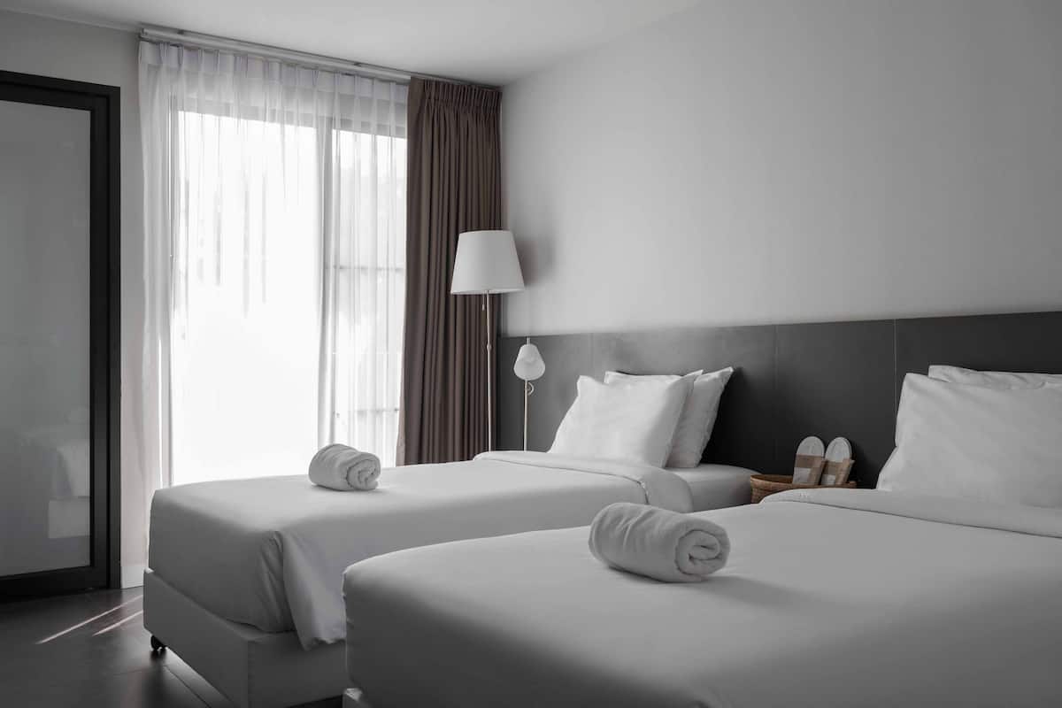 A hotel room with two beds, a large window with sheer curtains, a bedside lamp, and a wall-mounted TV.