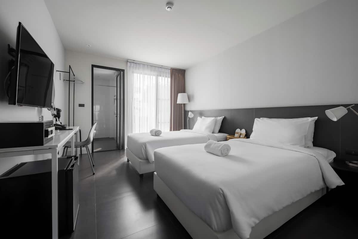 A modern hotel room with two beds, a desk, a chair, a TV, and a shower area.