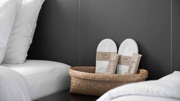 A neatly made bed with white linens, a dark wall, and a woven basket containing two white slippers and two brown paper bags with the text "POR".