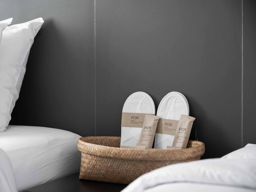 A neatly made bed with white linens, a dark wall, and a woven basket containing two white slippers and two brown paper bags with the text "POR".