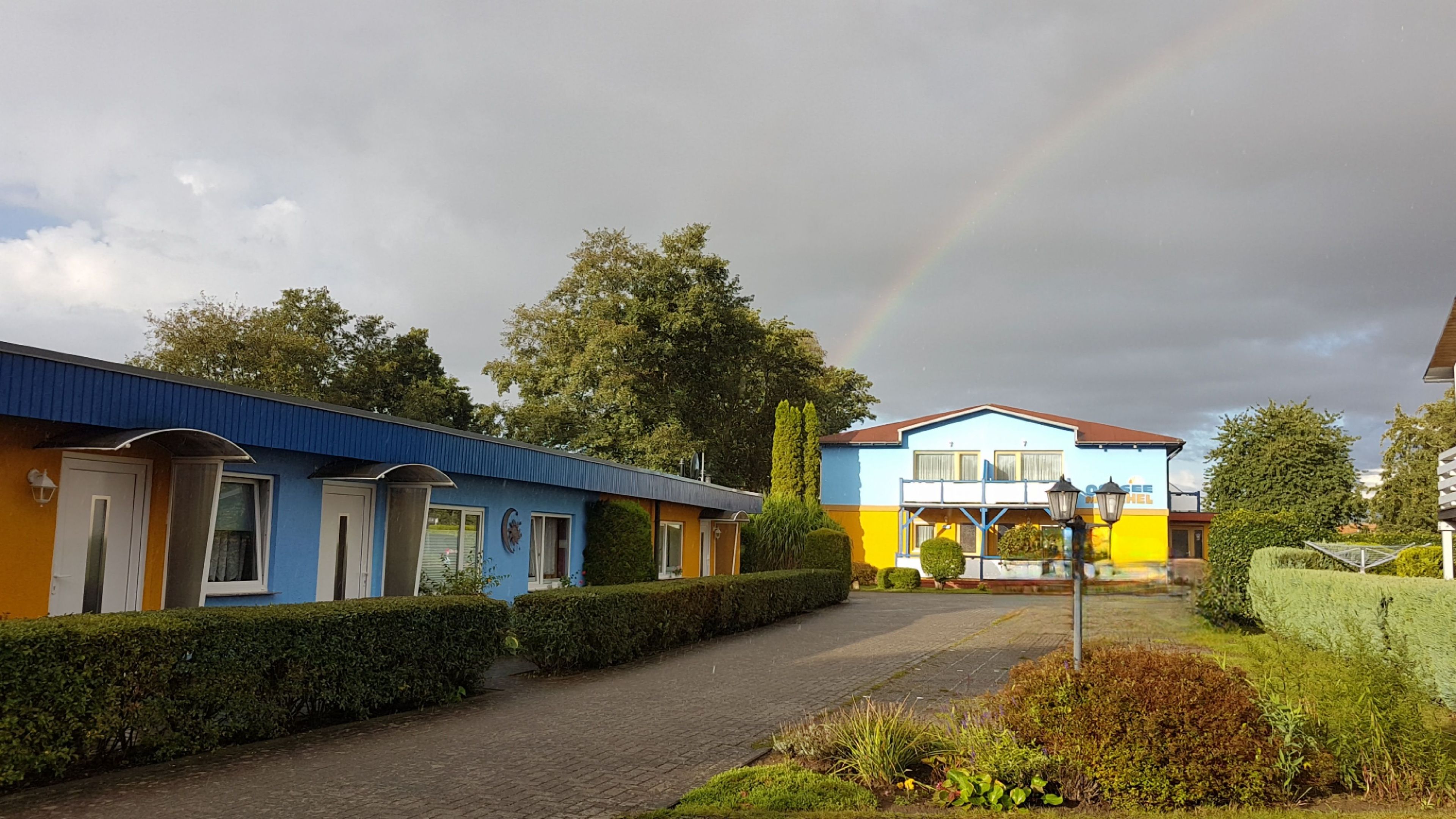 Eine Reihe bunter Gebäude mit einem gepflasterten Weg, einem Regenbogen in der Ferne und gepflegter Bepflanzung.