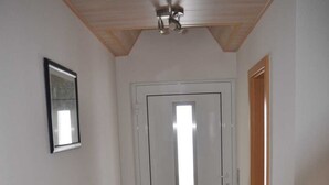 Ein Flur mit Fliesenboden, einer Tür mit Fenster, einem Deckenventilator und einem Lüftungsgitter an der Wand.