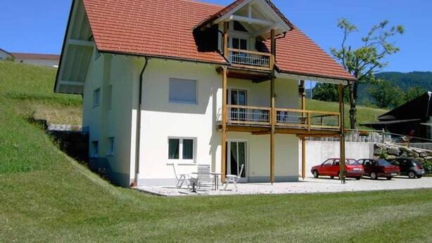 Ein zweistöckiges Haus mit rotem Ziegeldach, einem Balkon und einer kleinen angebauten Garage.