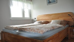 Ein Doppelbett mit Holz Kopfteil, ein Nachttisch mit Lampe und ein Fenster mit transparenten Vorhängen.