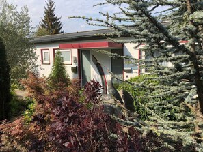 A small building with a red door and windows, surrounded by various plants and trees.