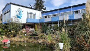 Un bâtiment moderne à la façade bleue et blanche, avec un étang au premier plan et un oiseau perché sur le toit.
