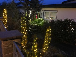 A residential area with houses, a stone wall, and Christmas lights on trees and bushes.
