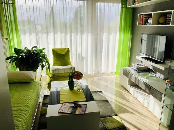A modern living room with a green armchair, a sofa, a coffee table with a book, a TV, and a bookshelf.