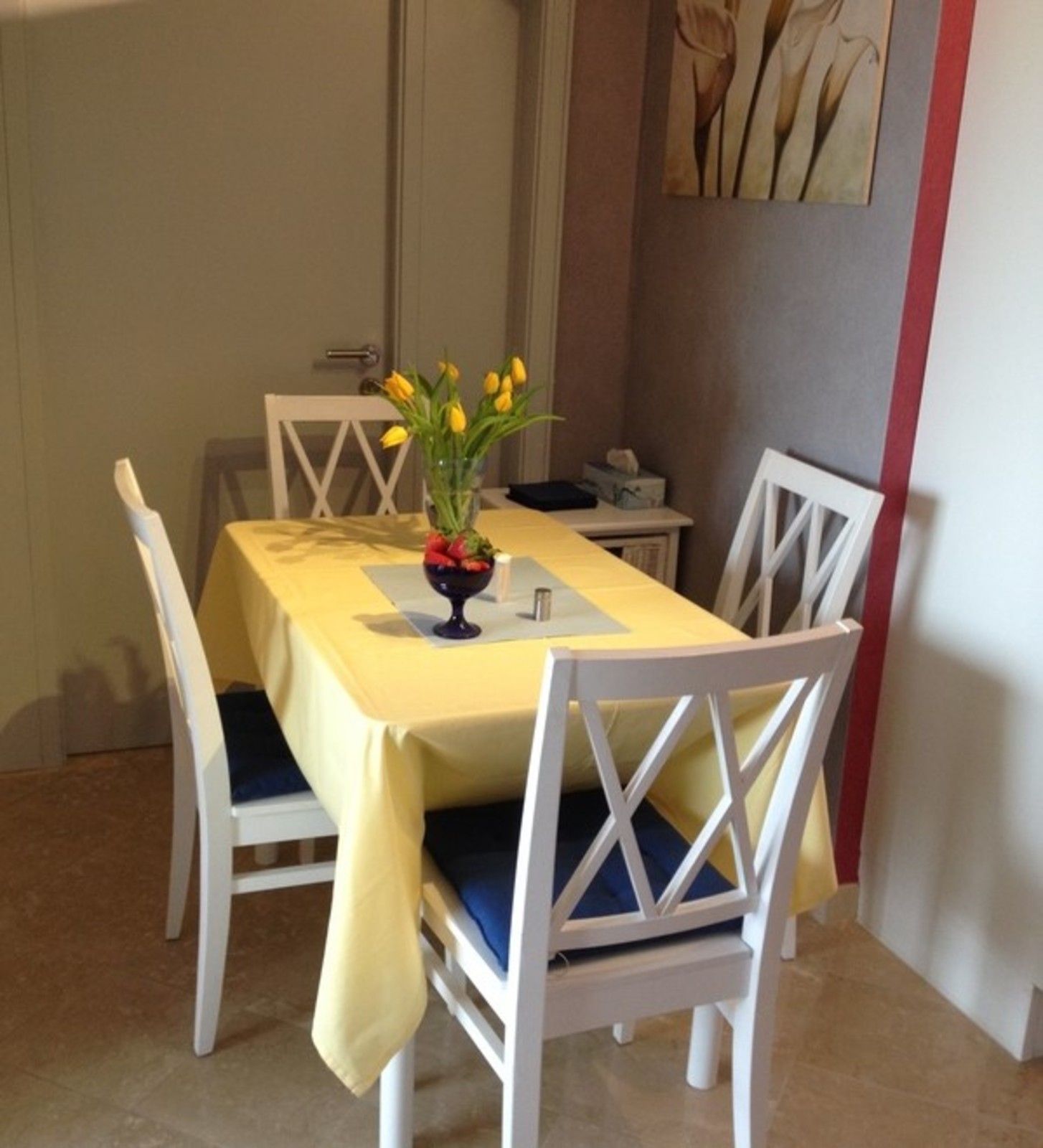 Ein Esstisch mit gelber Tischdecke, weiße Stühle mit blauen Kissen und eine Vase mit gelben Blumen.