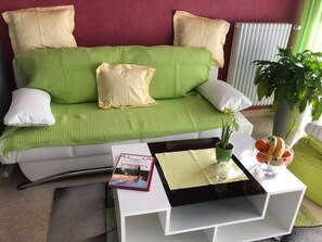 A green sofa with white and beige cushions, a white side table with a magazine, a glass of water, and a potted plant.