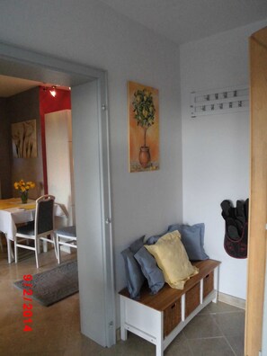 Ein Zimmer mit einer weißen Bank, einem Pflanzenbild, einem Kleiderständer und einem Essbereich mit Tisch und Stühlen.