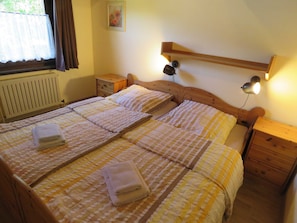 A double bed with two pillows and two folded towels. There is a wooden headboard, a nightstand with a lamp, and a small wooden drawer. A radiator is visible on the wall, and a window with curtains is on the left side. - Vacation home 87 Scout 42sqm for max. 4 persons with pet - Holiday home Scout 42 (Hollern-Twielenfleth)